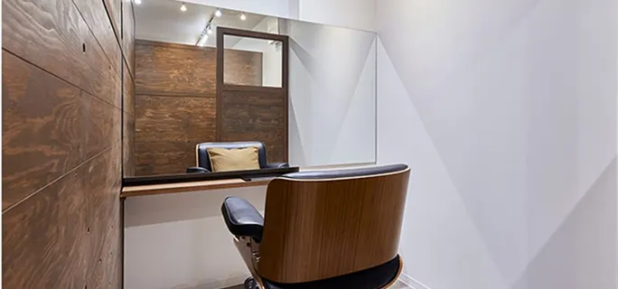 木目調の壁と大きな鏡が特徴の個室のヘアサロンブース。シンプルなデザインの木製チェアが鏡の前に置かれ、奥の鏡にクッション付きのチェアが映り込んでいる。