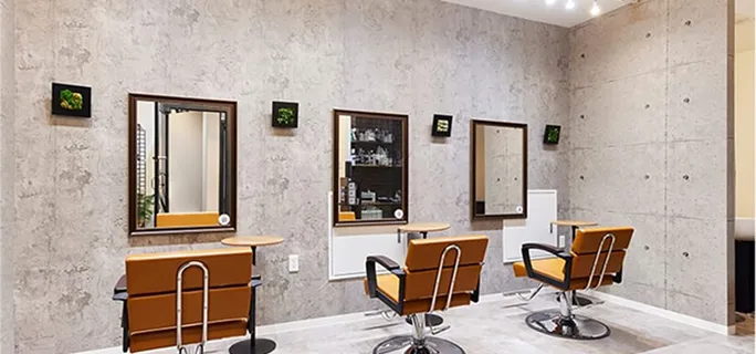 コンクリート調の壁に鏡が3枚並んだヘアサロンのセット面。鏡の下には木製のカウンターがあり、前にはキャメル色の椅子が設置されている。壁には小さな観葉植物のフレームが装飾され、天井にはライトが複数配置されている。