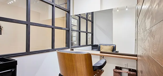 黒い格子状のガラスパーテーションが特徴の個室ヘアサロンブース。木製の壁と鏡があり、鏡の前には木目調のチェアが設置されている。足元にはブラウンの収納バッグがかけられている。