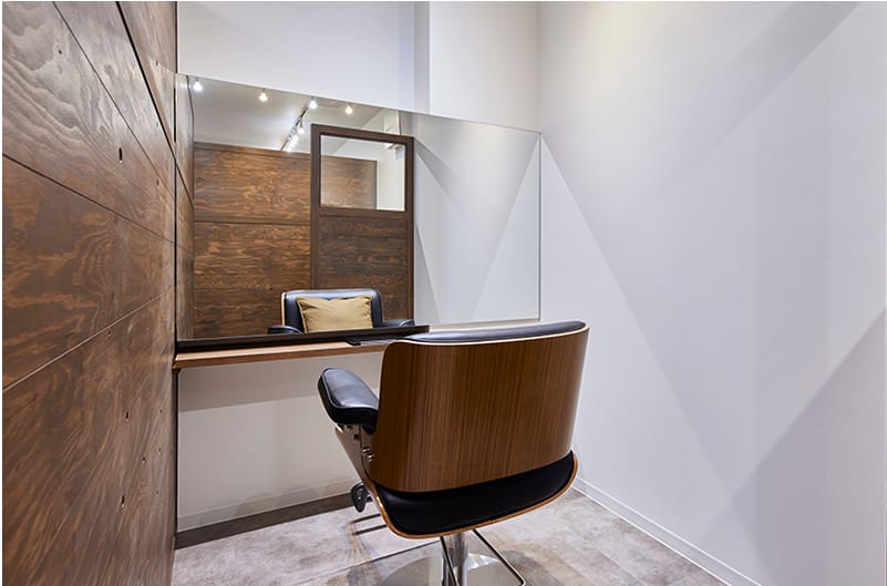 木目調の壁と大きな鏡が特徴の個室のヘアサロンブース。シンプルなデザインの木製チェアが鏡の前に置かれ、奥の鏡にクッション付きのチェアが映り込んでいる。